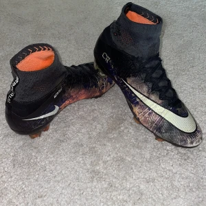 Nike Mercurial Superfly fotbollsskor - Säljer ett par NIKE MERCURIAL SUPERFLY CR7 SAVAGE BEAUTY FG fotbollsskor med högt skaft i svart, orange och lila mönster. Skorna har snörning, vit Nike-swoosh och textilmaterial med ACC-teknologi. Perfekta för dig som vill sticka ut på planen! På bild 4 och 5 ser ni små defekter samt de är inga sulor i men i helhet är skorna bra 👍. Skriv om du vill ha mer bilder eller har funderingar! Mvh Axel 