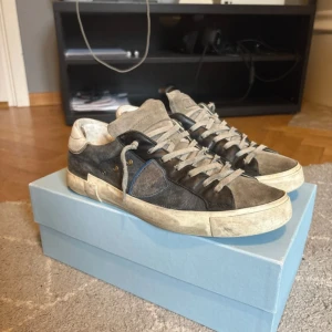 Philippe Model sneakers i mocka och skinn - Säljer ett par Philippe Model sneakers i svart och grå mocka med detaljer i skinn. Skorna har snörning, rund tå och platt sula. Klassisk design med diskret blå detalj på sidan och vita partier vid häl och sula.