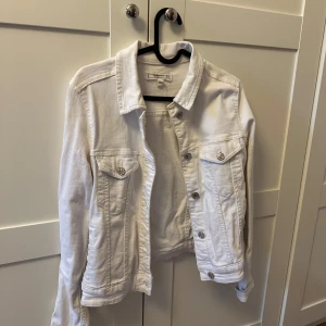 Vit jeansjacka från MANGO - Snygg vit jeansjacka från MANGO med klassisk krage och knappar framtill. Jackan har två bröstfickor med lock och knapp samt dekorativa sömmar. Perfekt för att lyfta din outfit med en fräsch och stilren look.