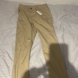 Beiga chinos från Kappahl, stl 164 - Snygga beiga chinos från Kappahl i storlek 164. Klassisk rak passform med fickor fram och bak, bälteshällor och knappstängning. Tillverkade i mjuk bomull som känns skön mot huden. Perfekta för en clean och stilren look. Helt nya, nypris 350kr