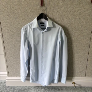 Ljusblå skjorta från BOSS - Säljer en stilren ljusblå skjorta från BOSS med klassisk krage och knäppning framtill. Skjortan har lång ärm och är i slim fit-modell, perfekt för en clean och dressad look. Materialet är lättskött och easy iron. Parfekt till sommaren!
