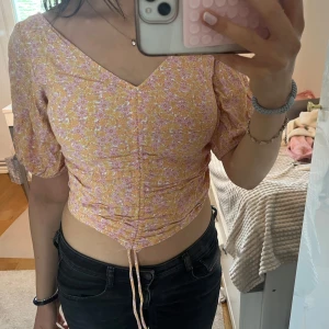 Blommig croppad topp med knyt - Superfin blommig croppad topp från H&M i ljusgul och lila med v-ringning och puffärmar. Toppen har knyt framtill och smockad rygg för extra komfort. Perfekt för vår och sommar!