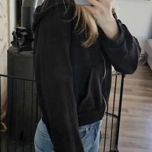 Svart hoodie med dragkedja från Lager 157 - Svart hoodie med dragkedja från Lager 157. Modellen har huva, ribbade muddar och fickor framtill. Tillverkad i en mjuk bomulls- och polyestermix. Perfekt för en avslappnad stil.