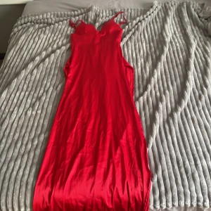 Röd långklänning i satin från Dennis Maglic - Säljer en röd långklänning i satin från Dennis Maglic. Klänningen har smala axelband, figurnära passform och djup urringning. Perfekt för dig som vill sticka ut med en elegant och stilren look.