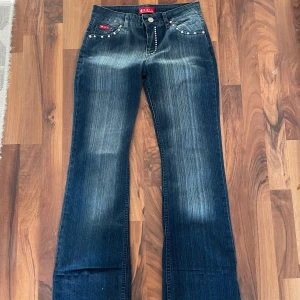 Mörkblå bootcut jeans med nitar - Säljer ett par mörkblå bootcut jeans med snygga slitningar och dekorativa nitar vid fickorna. Jeansen har normal passform och är tillverkade i bomull med lite stretch för extra komfort. Uppsydda. 