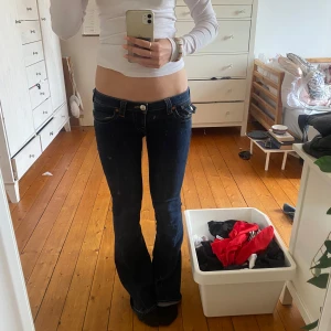Mörkblå lågmidjade jeans från True Religion - Säljer ett par mörkblå jeans från True Religion med låg midja och bootcut. Jeansen har dekorativa sömmar, fickor med lock och knapp bak samt slitna detaljer vid benslut. Klassisk True Religion-lapp bak i vitt och rött. De är i storlek 24 men passar några storlekar större, då de är stretchiga!