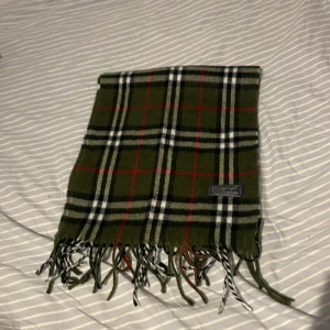 Grön rutig halsduk från Burberrys - Snygg grön rutig halsduk från Burberrys of London i 100% lammull. Klassisk design med röda, svarta och vita rutor samt fransar i ändarna. Perfekt accessoar för att lyfta din outfit med en tidlös brittisk touch.