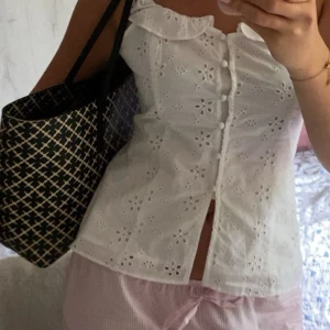 Vit broderad blus med volang - Superfin vit blus med broderade blommor och volang vid halsen. Knappar framtill och lätt croppad passform. Perfekt för sommarens alla tillfällen, riktigt söt och luftig med ärmlös design och romantiska detaljer.