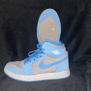 Nike Air Jordan 1 blå och grå sneakers - Nike Air Jordan 1 sneakers i en snygg kombination av ljusblått och grått. Skorna har klassisk hög siluett, blå snörning och vit sula med blå detaljer. Ovandelen är i mocka och textil, med ikoniska Air Jordan-loggan på sidan. Perfekta för dig som vill sticka ut med stil.