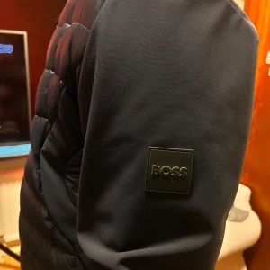 Svart pufferjacka från BOSS - Snygg svart pufferjacka från BOSS med quiltad framsida och släta ärmar. Jackan har en grön dragkedja som ger en cool kontrast och BOSS-logga på ärmen. Lättviktig och vattenavvisande, perfekt för höst och vinter. Fodrad med dun för extra värme.