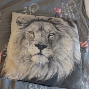 Svart prydnadskudde med lejonmotiv - Pryddnadskudde med ett stort, detaljerat lejonansikte tryckt framtill. Perfekt för dig som vill sticka ut med ett djurmotiv.