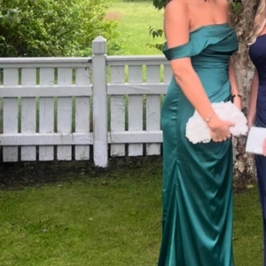 Grön axelbandslös långklänning - Säljer en elegant smaragdgrön långklänning med offshoulder-design och figurnära passform. Klänningen har en vacker drapering över bysten och släpper mjukt längs kroppen. Perfekt för bal eller fest.