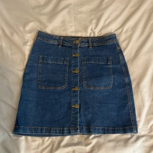 Jeanskjol med knappar framtill - Säljer en klassisk blå jeanskjol med två stora fickor framtill och snygga metallknappar längs hela framsidan. Kjolen har bälteshällor och är i en rak, kort modell som passar perfekt till vardagslooken.