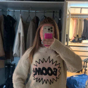 Beige fluffig tröja med BOOM-tryck - Säljer nu min populära BOOM tröja. Supermjuk och fluffig beige tröja med ett stort BOOM-tryck i svart och rosa framtill. Tröjan har lång ärm och en avslappnad passform, perfekt för dig som gillar statement-plagg och vill sticka ut lite extra. 