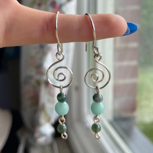 Silvriga örhängen med gröna pärlor - Unika handgjorda örhängen i silverfärgad metall med spiralformad design. Hängande detaljer med tre pärlor i olika gröna nyanser, där mittenpärlan är ljusare mintgrön. Perfekta för dig som gillar boho eller naturinspirerad stil.