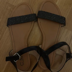 Svarta sandaler med glitterdetalj - Stilrena sandaler från C'M Paris med svarta remmar och glittrig detalj framtill. Snyggt spänne vid ankeln och platt sula för en bekväm känsla. Perfekta för varma dagar när du vill ha något enkelt men ändå glammigt.