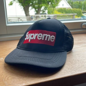 Supreme svart truckerkeps med mesh - Svart truckerkeps från Supreme med klassisk röd box logo framtill i broderad vit text. Kepsen har böjd skärm och mesh på sidorna och bak för extra ventilation. Justerbar passform baktill. Perfekt för dig som vill ha streetstyle med ikonisk känsla.