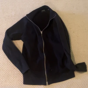 Svart cardigan tröja med dragkedja - Svart cardigan jacka från Collage med hel dragkedja framtill. Jackan har en enkel och stilren design med långärmade ärmar och hög krage. Perfekt för lager-på-lager och passar till många olika outfits.