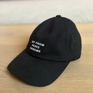 Svart keps Drôle de Monsieur - Svart keps från Drôle de Monsieur med vit broderad text 'NOT FROM PARIS MADAME' framtill. Justerbar rem baktill med metallspänne. Tillverkad i 100% bomull för en klassisk och clean look. Helt oanvänd. Nypris 850kr.