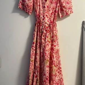 Blommig långklänning från H&M XS - Superfin långklänning från H&M i blommigt mönster med rosa, röda och gröna toner. Klänningen har korta puffärmar, knappar framtill och ett knytband i midjan. Tillverkad i tunn bomull, perfekt för varma dagar och somriga vibes.