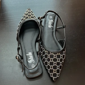 Svarta ballerinaskor med strass från Shein - Svarta ballerinaskor från Shein med spetsig tå och tunna remmar. Skorna har ett glittrigt mönster av små strass-stenar i ett rutigt nät över hela ovandelen. Platt sula och öppen häl ger en luftig känsla. Perfekta för dig som gillar att sticka ut.