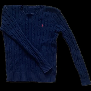 Mörkblå stickad tröja från Ralph Lauren - Kabelstickad mörkblå tröja från Ralph Lauren med klassisk rund halsringning och röd broderad logga på bröstet. Tröjan har lång ärm och ribbade muddar vid ärmslut och nederkant. Perfekt för en stilren och tidlös look.