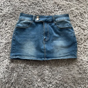 Blå jeanskjol med dubbla knappar - Snygg blå jeanskjol med klassisk femficksdesign och dubbla silverfärgade knappar i midjan. Kjolen är kort och har bälteshällor samt kontrastsömmar. Perfekt för dig som gillar en enkel men trendig denimlook.                                       Köpte den här kjolen begagnad för 200 kr, men den var tyvärr för stor och har aldrig blivit använd. Hoppas någon annan kan få glädje av den!
