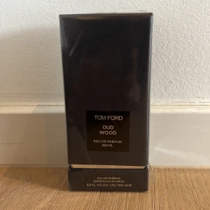 Tom Ford Oud Wood EdP 100ml - Tom Ford Oud Wood Eau de Parfum 100 ml i elegant svart kartong med diskret mönster. Lyxig och exklusiv doft med träiga och orientaliska toner. Förpackningen är rektangulär och tillverkad av kartong med plastfilm runt.