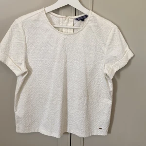 Vit strukturerad blus Tommy Hilfiger - Vit blus från Tommy Hilfiger med korta ärmar och rund hals. Tyget har en snygg strukturerad yta och en liten guldfärgad Hilfiger-detalj nertill. Blusen har dragkedja i ryggen för enkel påklädning. Perfekt för en clean och stilren look. Passar storlek S.