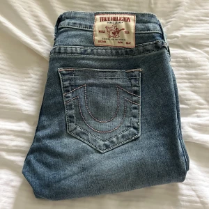 True Religion ljusblå bootcut jeans - Säljer ett par ikoniska ljusblå jeans från True Religion med klassiska kontrastsömmar och tydlig logga på bakfickan. Modellen har låg midja och bootcut-ben, vilket ger en snygg retrovibe. Perfekta för dig som gillar streetstyle och vill sticka ut.