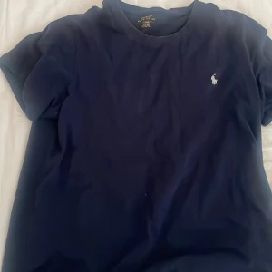 Mörkblå t-shirt från Polo Ralph Lauren - Klassisk mörkblå t-shirt från Polo Ralph Lauren med broderad vit logga på bröstet. T-shirten har rund halsringning och korta ärmar. Perfekt basic-plagg med snygg passform och stilren look.