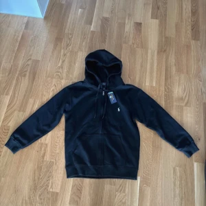 Svart hoodie från Polo Ralph Lauren - Svart hoodie från Polo Ralph Lauren med klassisk broderad logga på bröstet. Tröjan har dragkedja framtill, huva med snörning och två fickor. Tillverkad i mjuk bomull, perfekt för en chill och stilren look. Den är helt ny!