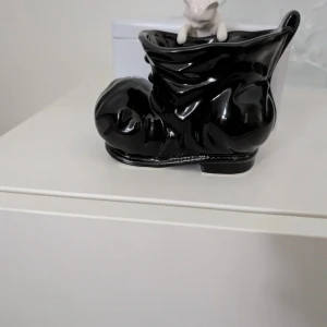 Svart keramikstövel med katt - Dekorativ stövel i svart keramik med glansig finish och en vit katt som klättrar över kanten. Stöveln har rund tå och detaljerad form, perfekt som prydnad eller liten förvaring.