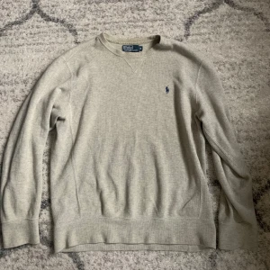 Grå tröja från Polo Ralph Lauren - Snygg grå tröja från Polo Ralph Lauren med klassisk rund halsringning och broderad blå logga på bröstet. Tröjan är långärmad och har ribbade muddar vid ärmslut och nederkant. Perfekt basic att ha i garderoben.