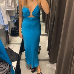 Blå långklänning - Säljer en snygg blå långklänning med smala axelband. Klänningen har en v-ringad front med twistad detalj och metallring, samt cut-out under bysten. Materialet är glansigt. Perfekt sommarklänning/midsommarklänning. Använd en gång bara!