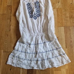 Vit kortklänning med blå brodyr - Superfin vit kortklänning från Gina Tricot med långa ärmar och blå broderade detaljer framtill. Klänningen har knytning i halsen, resår i midjan och tunna blå ränder nertill. Perfekt för sommarens varma dagar och har en bohemisk vibe.