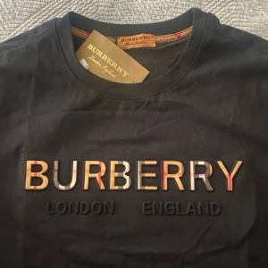 Svart Burbery t shirt  - Svart t-shirt från Burbery med klassisk rund hals och korta ärmar. Stor broderad Burberry-logga framtill i beige, rött och svart med rutiga detaljer. Tillverkad i mjuk bomull för en skön känsla. Perfekt för dig som gillar stilrena designer med lyxig touch.
