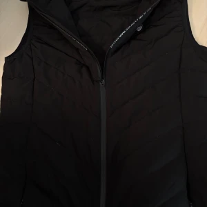 Svart dunväst från Emporio Armani EA7 - Snygg svart dunväst från Emporio Armani EA7 med quiltad design och dragkedja framtill. Västen har en stilren look med EA7-logga på bröstet och diskreta fickor i sidorna. Perfekt för lager-på-lager och streetwear-vibe.