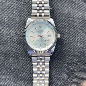 silverfärgad klocka - Snygg klocka med klassisk jubilee-länk i silverfärgat stål. Urtavlan är ljus med datumfönster och tydliga indexmarkeringar. Boetten har räfflad kant och armbandet har den ikoniska kronan på spännet. En tidlös accessoar med exklusiv känsla.