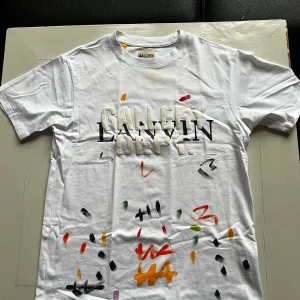 Vit Lanvin x Gallery Dept. t-shirt med tryck - Vit t-shirt från Lanvin x Gallery Dept. med coolt tryck framtill och bak, samt färgstänk i olika nyanser. Klassisk passform med korta ärmar och rund hals. Materialet är mjuk bomull som känns skönt mot huden. Perfekt för dig som gillar streetwear och unika detaljer.