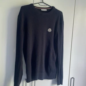 Svart stickad tröja från Moncler - Svart stickad tröja från Moncler med diskret logga på bröstet. Tröjan har rund halsringning, ribbade muddar och långärmad passform. Perfekt för dig som gillar stilrena och exklusiva plagg med en klassisk vibe.