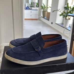 Mörkblå mockaloafers - Snygga mörkblå loafers i mocka från HAMMER STEIN. Skorna har en vit platt sula och en dekorativ rem med metallspänne över vristen. Insidan är fodrad i brunt äkta läder för extra komfort. Perfekta för en stilren och avslappnad look. Ny pris 3200kr