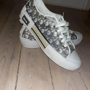 Dior sneakers med monogrammönster - Snygga sneakers från Dior med klassiskt monogrammönster i grått och vitt. Skorna har vita snören, rund tå och platt sula. Sidan pryds av en beige och svart detalj samt Dior-logga. Perfekta för dig som gillar exklusiv streetstyle.