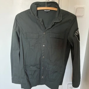 Grön fieldjacket från Pomp De Lux - En tunn, stilren fieldjacket som är grön i färgen. Perfekt för svala sommarkvällar eller för en promenad på stan, med tryck i ryggen och på axeln är den unik och säljs inte längre i butik. Storleken är 158 - 164. Skriv i PM om ni har frågor😊