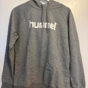 Grå hoodie från Hummel med tryck - Snygg grå hoodie från Hummel med vit logga framtill. Tröjan har huva med snörning och långa ärmar. Perfekt för chill dagar eller när du vill ha en sportig look. Mjuk och skön i bomullsblandning.