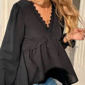 Svart blus med spets och v-ringning - Supersöt svart blus med v-ringad hals och dekorativ spetskant. Blusen har lång ärm och en lös, flowy passform som ger en chill vibe. Perfekt att styla med jeans eller kjol för en trendig look.