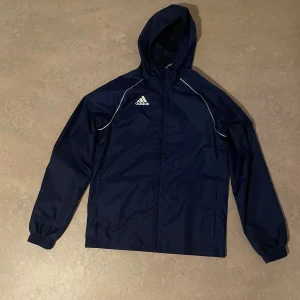 Mörkblå vindjacka från Adidas - Snygg mörkblå vindjacka från Adidas med huva och vita detaljer längs axlarna. Jackan har dragkedja framtill, elastiska muddar och klassisk Adidas-logga på bröstet. Perfekt för blåsiga dagar och sportiga outfits. Användes knappt en höst.