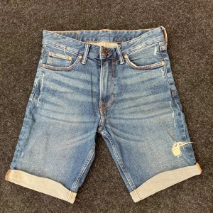 Blå jeansshorts HM - Snygga blå jeansshorts från HM med klassisk femficksdesign, uppvikta benslut och slitningar på ena benet för en cool look. Perfekta för sommardagar och enkla att matcha med t-shirt eller hoodie. Storlek 165