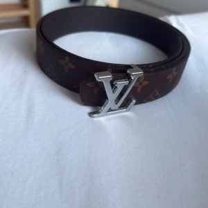 Brunt bälte  - Säljer ett brunt  bälte i skinn med klassiskt monogrammönster och silvermetall framtill. Bältet har exklusiv känsla och är perfekt för dig som vill sticka ut med en ikonisk accessoar.