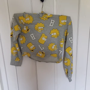 Grå Simpsons hoodie från H&M - Grå hoodie från H&M med färgglatt Simpsons-tryck av Bart och Lisa över hela tröjan. Långärmad och med huva, perfekt för dig som älskar tecknade serier och vill sticka ut. Mjuk bomullsmix som är skön att ha på sig. Snygg och lekfull stil!